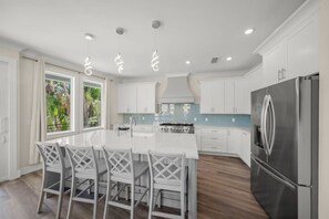 Cocina moderna con armarios blancos, una isla central con taburetes y un refrigerador de acero inoxidable, con vistas a zonas verdes.