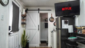 A modern kitchen with a large flat-screen TV displaying 'NETFLIX,' a door with a black towel, and a potted plant. - Cozy Container, HotTub, King Bed, Pets, Wifi, 1 Night Stays, Griddle, Firepit (Chillicothe)