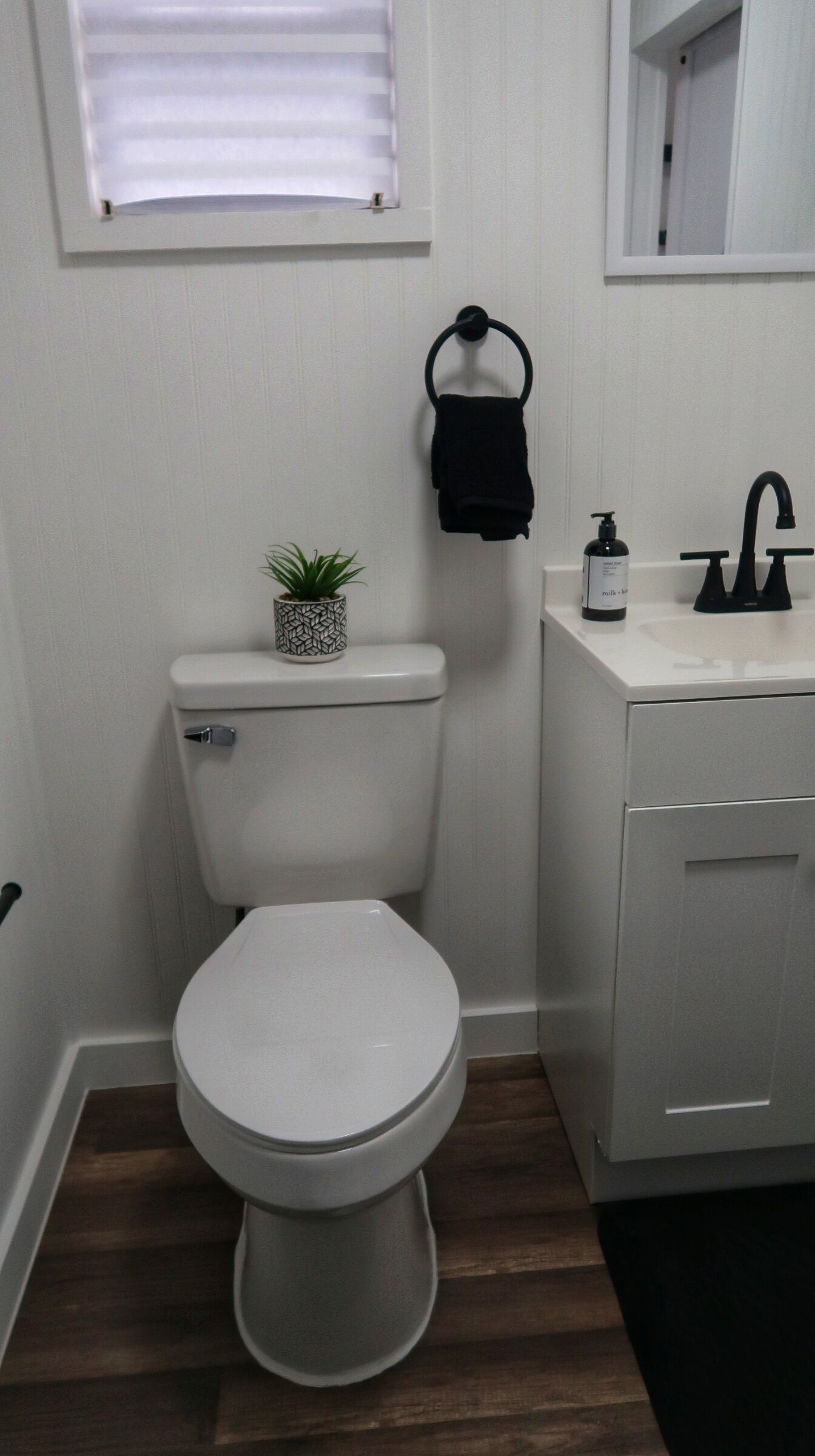 A small, modern bathroom with a white toilet, a sink with a black faucet, a towel ring, and a small potted plant.