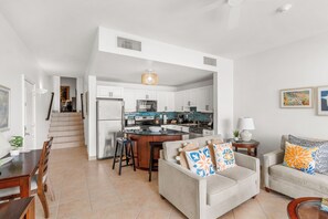 A modern kitchen with white cabinets, a central island, and stainless steel appliances. There is a dining area with a wooden table and chairs, and a living room with a sofa and a coffee table.