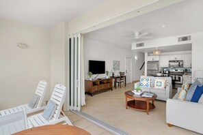 A spacious living area with a dining table, two white armchairs, a sofa, and a kitchen in the background.