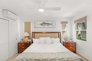 A bedroom with a bed, two bedside tables with lamps, a ceiling fan, a wall-mounted air conditioner, and a window with blinds.