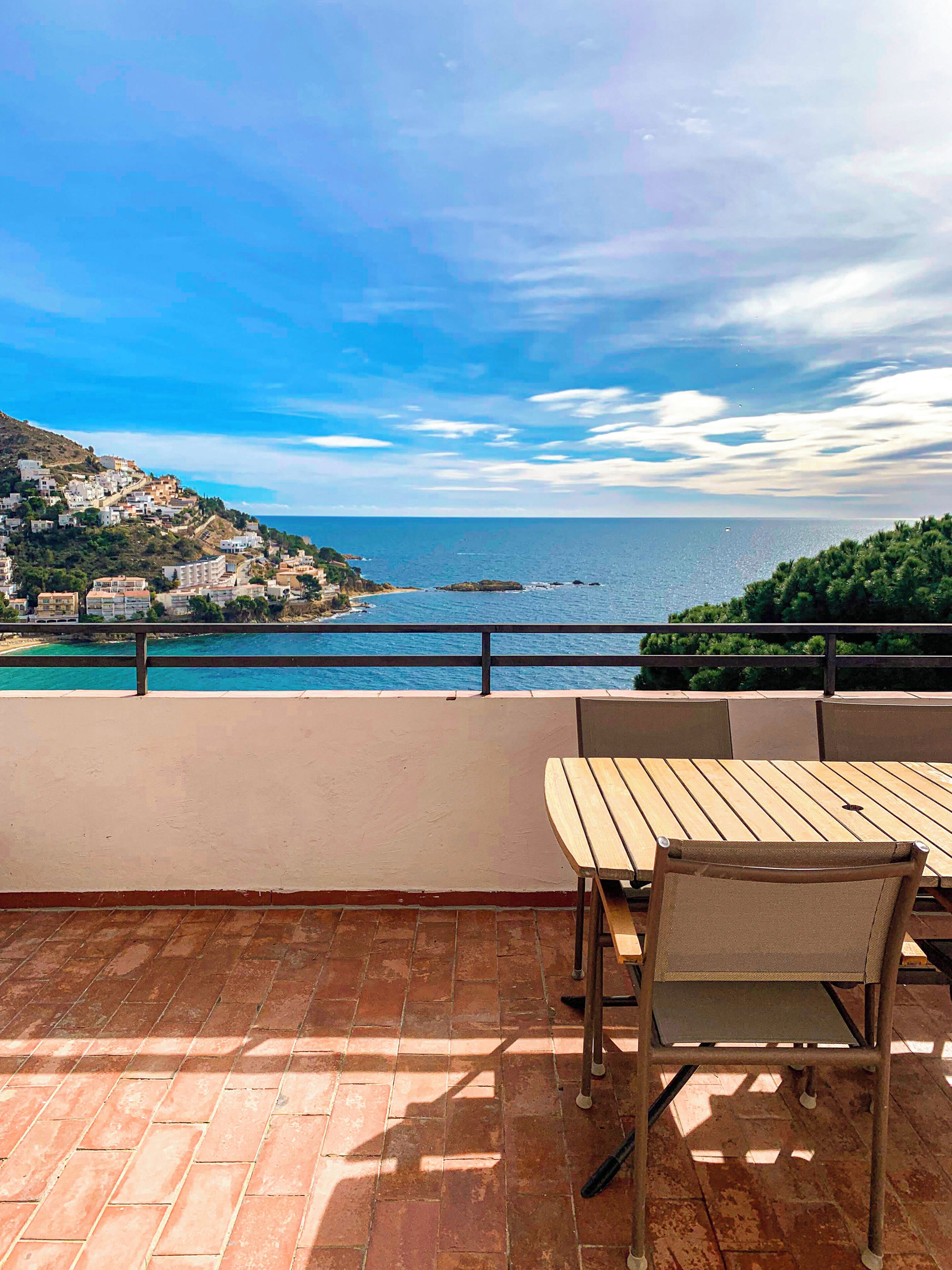 A terrace with a wooden table and chairs, overlooking a coastal town with buildings and the sea.