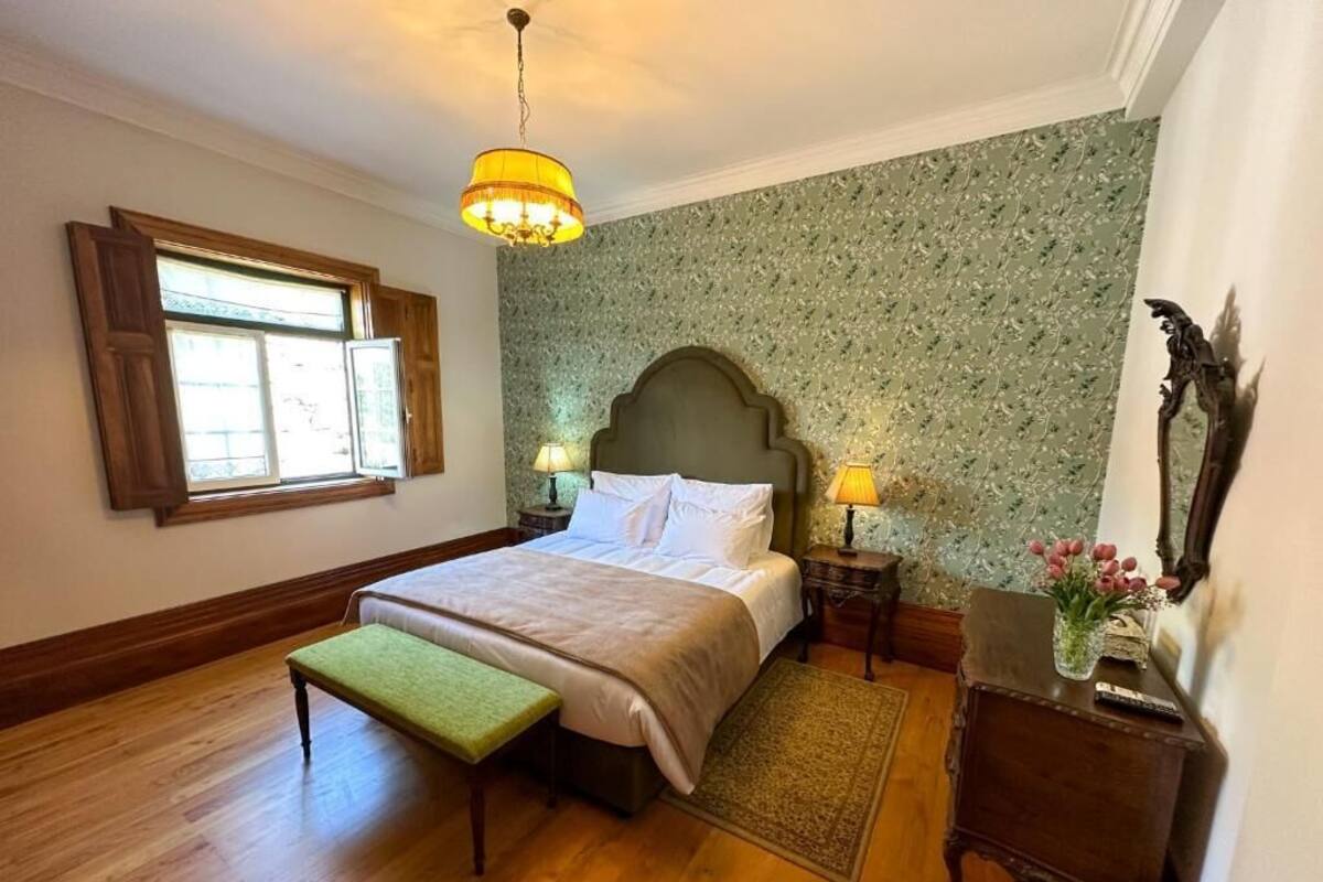 A bedroom with a large bed, wooden floor, a bench, a nightstand with a lamp, and a wall-mounted mirror.
