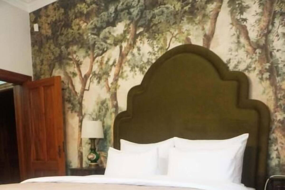 A neatly made bed with a green bench in front, a wooden headboard, and a wall with a tree mural.