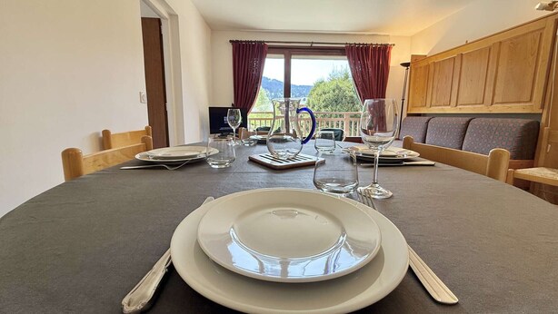 Une table à manger dressée pour quatre personnes, avec assiettes, couverts et verres, un téléviseur en arrière-plan et une vue sur l’extérieur par une fenêtre.