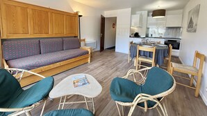 Un salon avec un canapé, deux fauteuils, une petite table et, au fond, une cuisine.