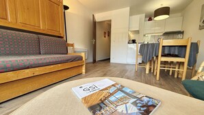 Un salon confortable avec un canapé, un magazine posé sur le sol, une table à manger avec des chaises, et une cuisine en arrière-plan.