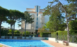 A modern building with a swimming pool, surrounded by trees and a fenced area. - Apartment With Pool in Lignano Pineta (Lignano Sabbiadoro)