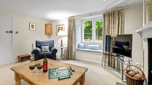 Un salón acogedor con un sillón azul, una mesa de centro de madera con copas de vino y un juego de mesa, un televisor y una ventana con vistas a zonas verdes.