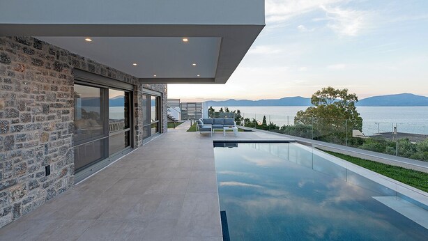 A modern outdoor pool area with a stone wall and glass doors, overlooking a body of water and distant mountains. - Design villa with immaculate views in Theologos (Theologos)