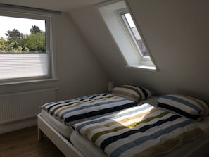 Et soveværelse med skråvæg, ovenlys, seng med stribet sengetøj og et vindue med persienner.