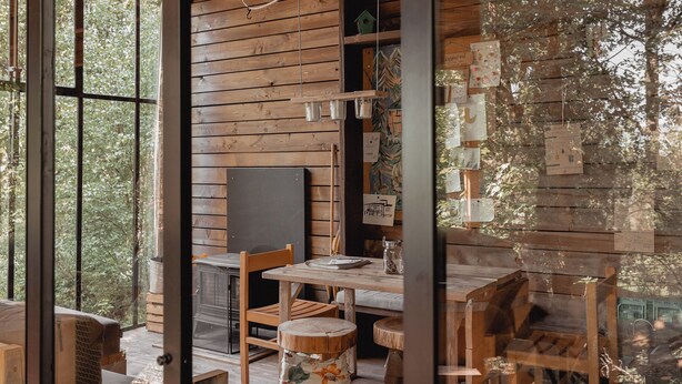A rustic cabin interior with wooden walls, a dining area, and a large window overlooking a forest. - Nutchel Cosy and Nature Cabin for 4 (Martelange)