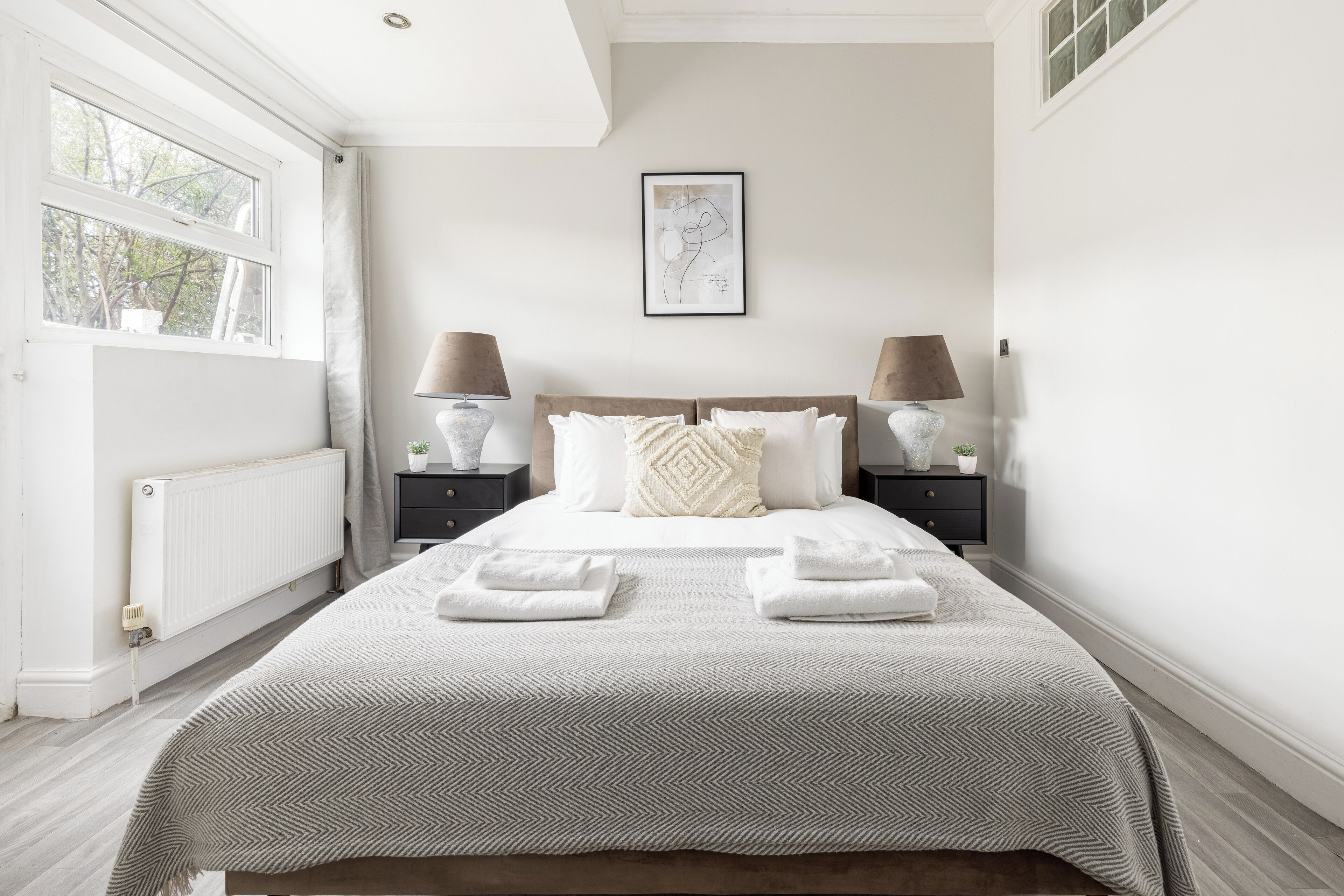 A bedroom with a large bed, two bedside tables with lamps, a radiator, and a window with curtains.