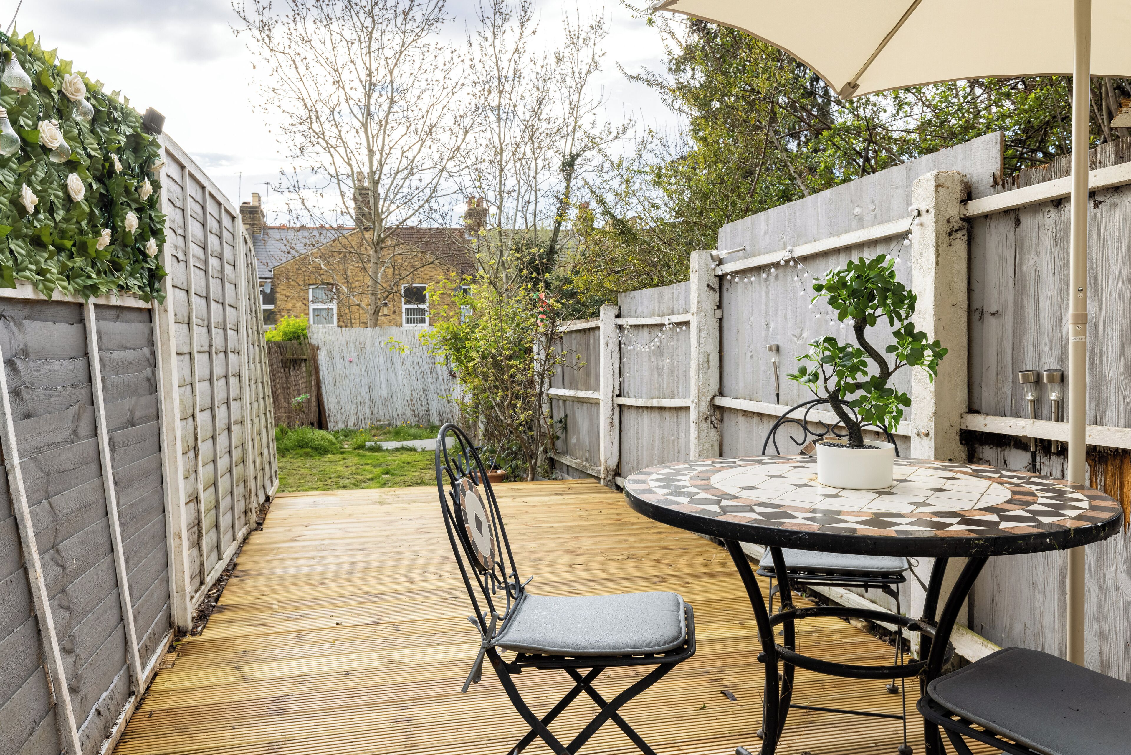 A wooden deck with a round table and chairs, surrounded by a wooden fence and a garden.
