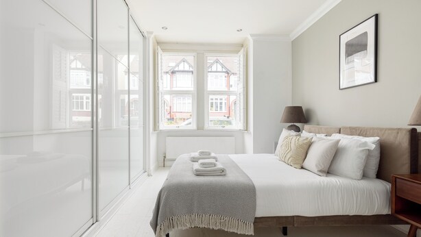 A bedroom with a large bed, a bedside table, a lamp, and a window with a view of houses.