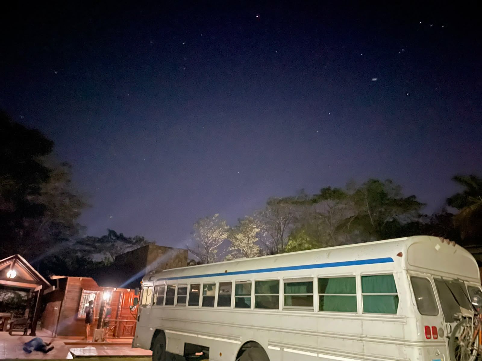 A white bus parked at night with a blue stripe, a building with a porch, and trees in the background.