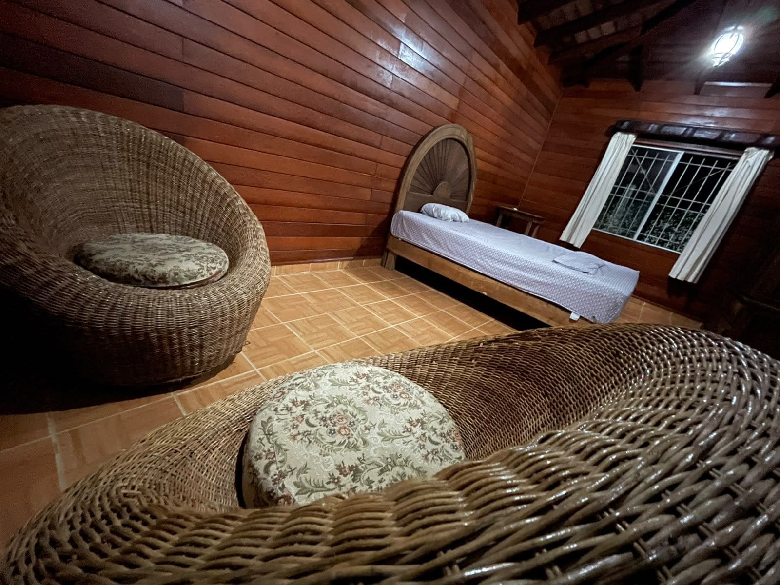 A room with a wooden ceiling, a bed with a white bedspread, and two wicker chairs with cushions.