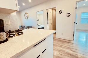 Una cocina con una encimera, una cafetera y frascos. Hay un reloj en la pared y una puerta que da a otra habitación.