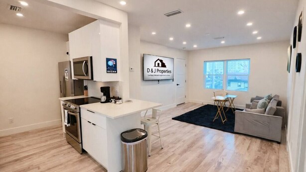 A modern kitchen with a central island, a dining area with a small table and chairs, a living room with a sofa, and a large window. - Daytona Double Delight (Daytona Beach)