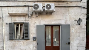 A building facade with a stone wall, a wooden door, and two air conditioning units mounted above the door. - Casa Angelina - Cozy stay near the most beautiful destinations in Puglia (Molfetta)
