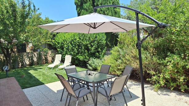 Eine Terrasse mit Glastisch und Stühlen, einem Sonnenschirm und einem Garten mit verschiedenen Pflanzen und einem Zaun.