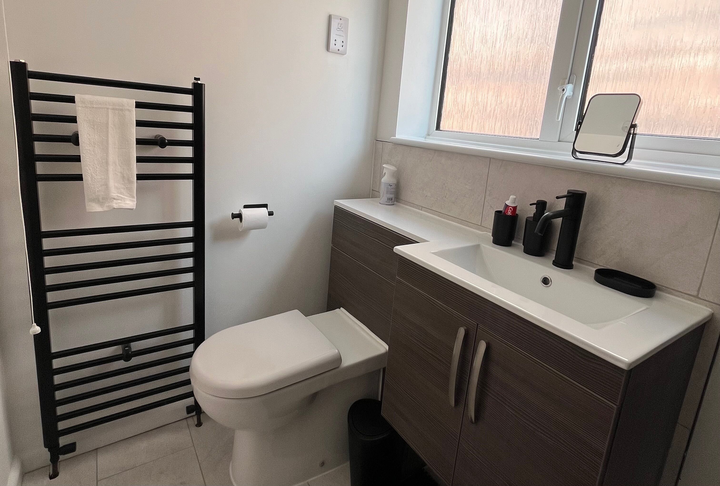 A bathroom with a black towel rack, a white toilet, a sink with a mirror, and a window.