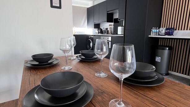 A modern kitchen with dark cabinets, a washing machine, and a dining area with black plates and wine glasses. - Nottingham 2-bed House, Sleeps 4, Pet Friendly (Nottingham)