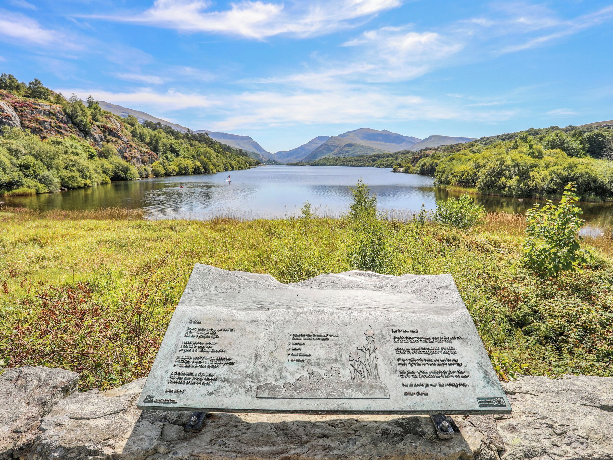 A stone sign with text and a map, placed on a rocky surface near a lake surrounded by hills.