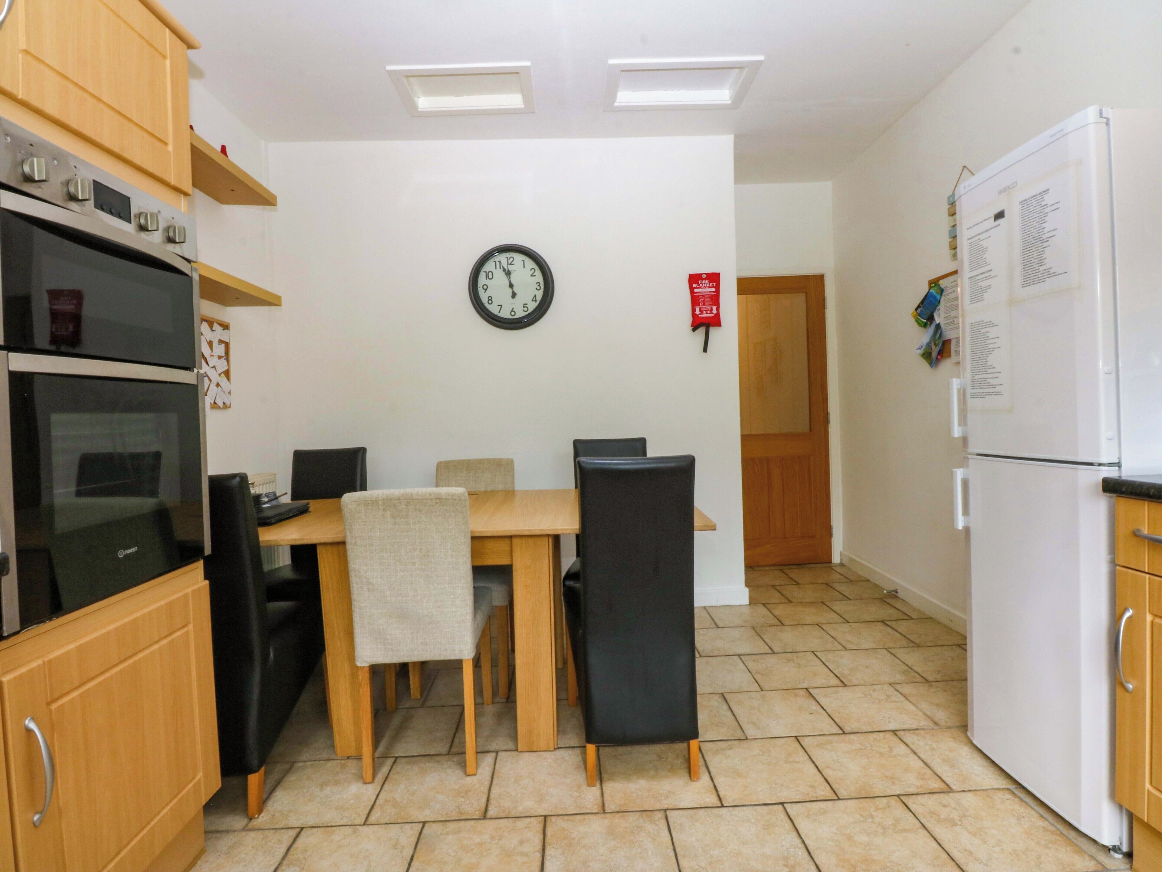 A compact kitchen with a dining table, chairs, a clock, and a refrigerator.