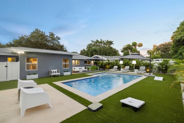 A modern backyard with a swimming pool, white lounge chairs, and a covered patio area.