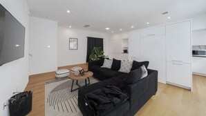 Een moderne woonkamer met een zwarte bank, een salontafel en een televisie. Er is een keuken met witte kasten en een eettafel.