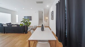 Een moderne woonkamer met een witte eettafel, zwarte gordijnen, een plant in pot en een bank.