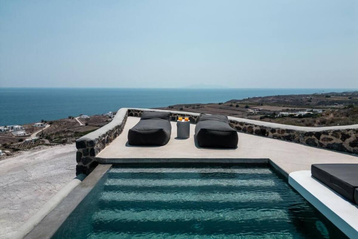 An infinity pool with black lounge chairs and a stone wall, overlooking a coastal landscape with buildings and a clear sky.