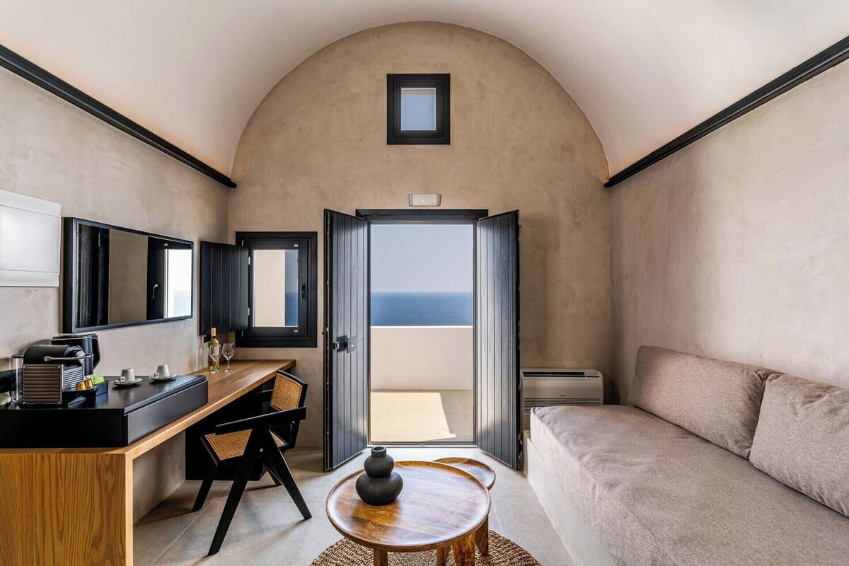 A compact room with a wooden desk, a sofa, a small table, and a view of the sea through an open door.