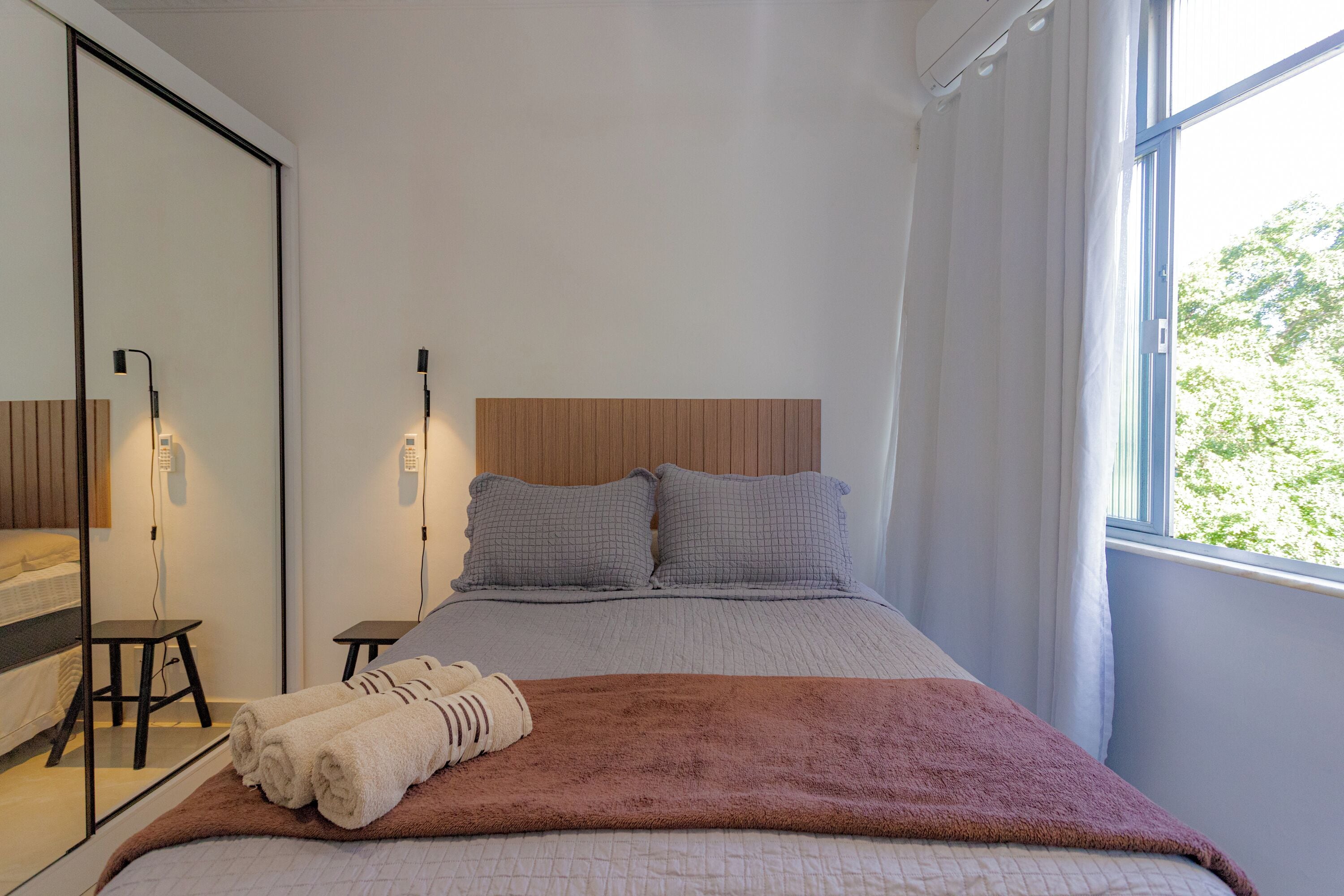 Un dormitorio con cama, espejo, mesita de noche y ventana con cortinas.