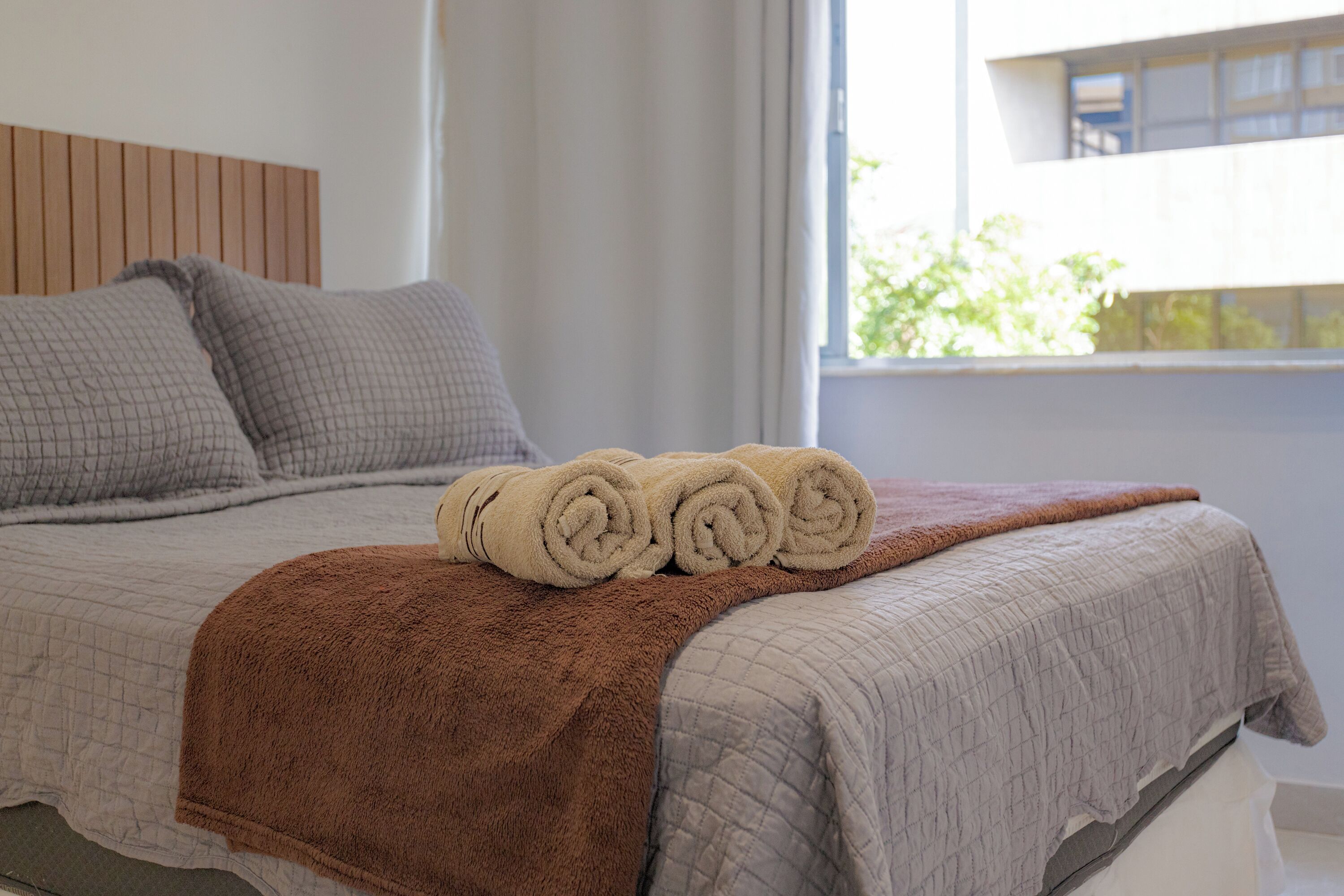 A neatly made bed with rolled towels and a brown blanket, a wooden headboard, and a window with a view of greenery outside.