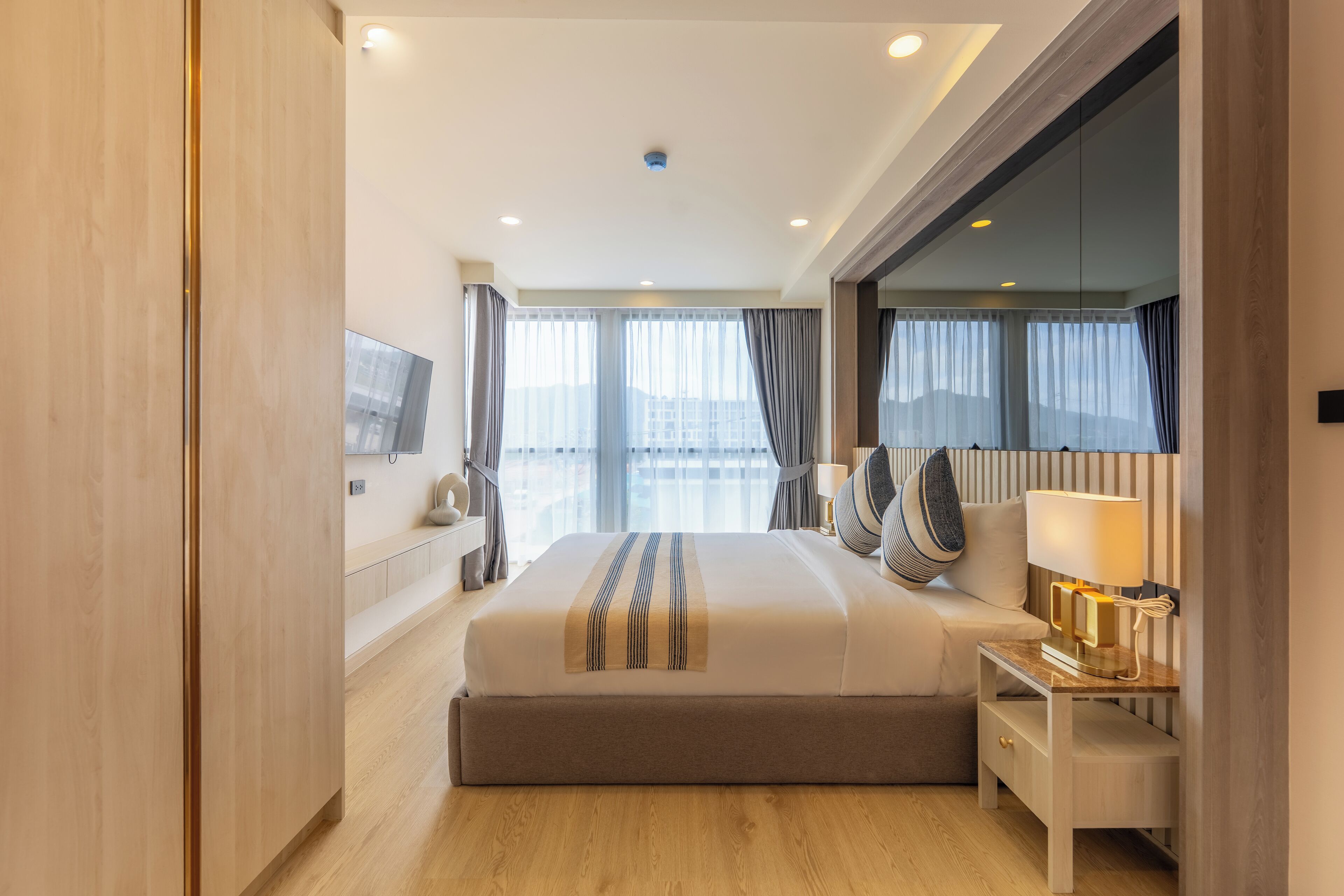 A modern hotel room with a large bed, bedside table with a lamp, and a view of the city through large windows.