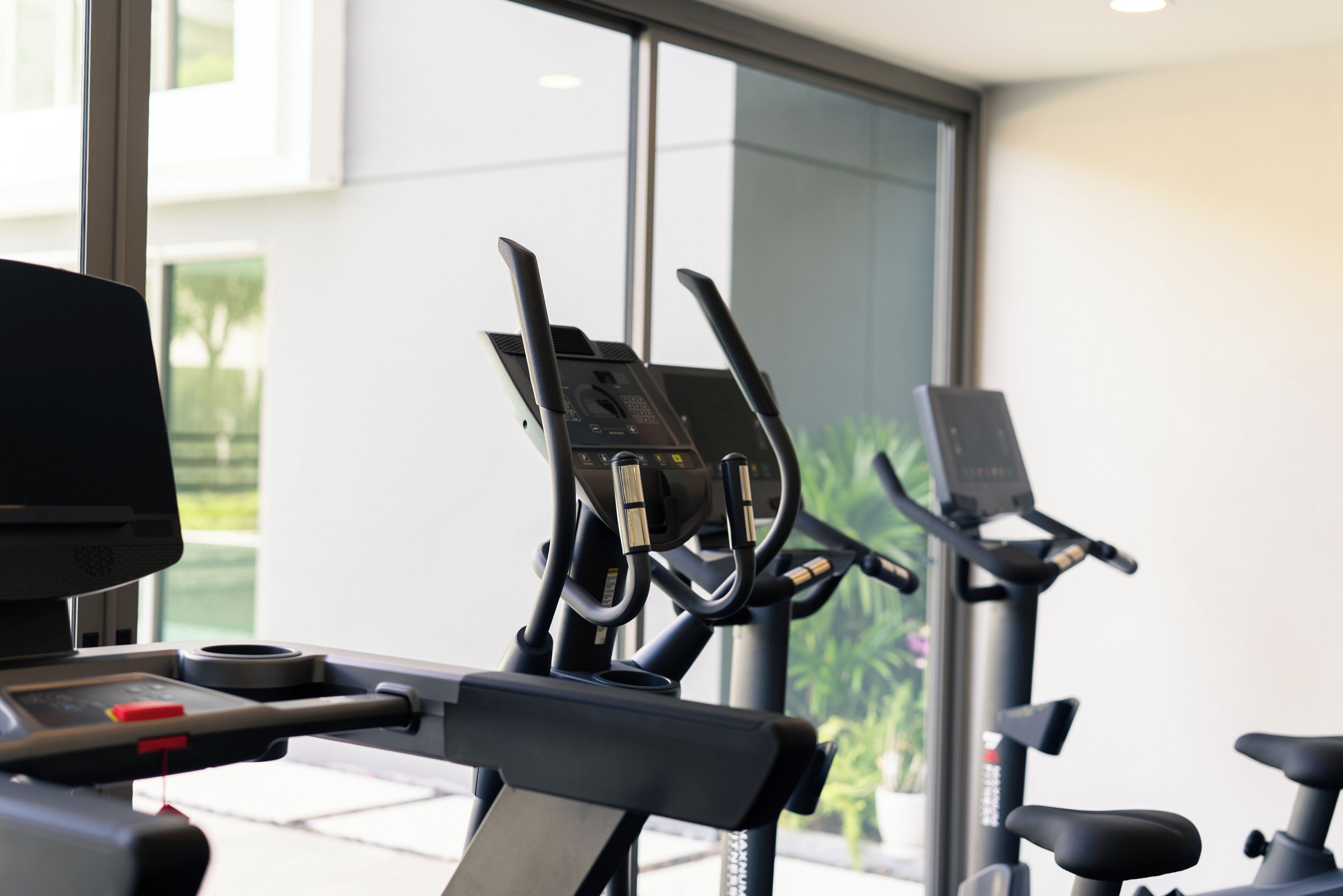 Un vélo d’appartement dans une salle de sport, doté d’un écran numérique, une fenêtre donnant sur un paysage verdoyant et un tapis de course à l’arrière-plan.