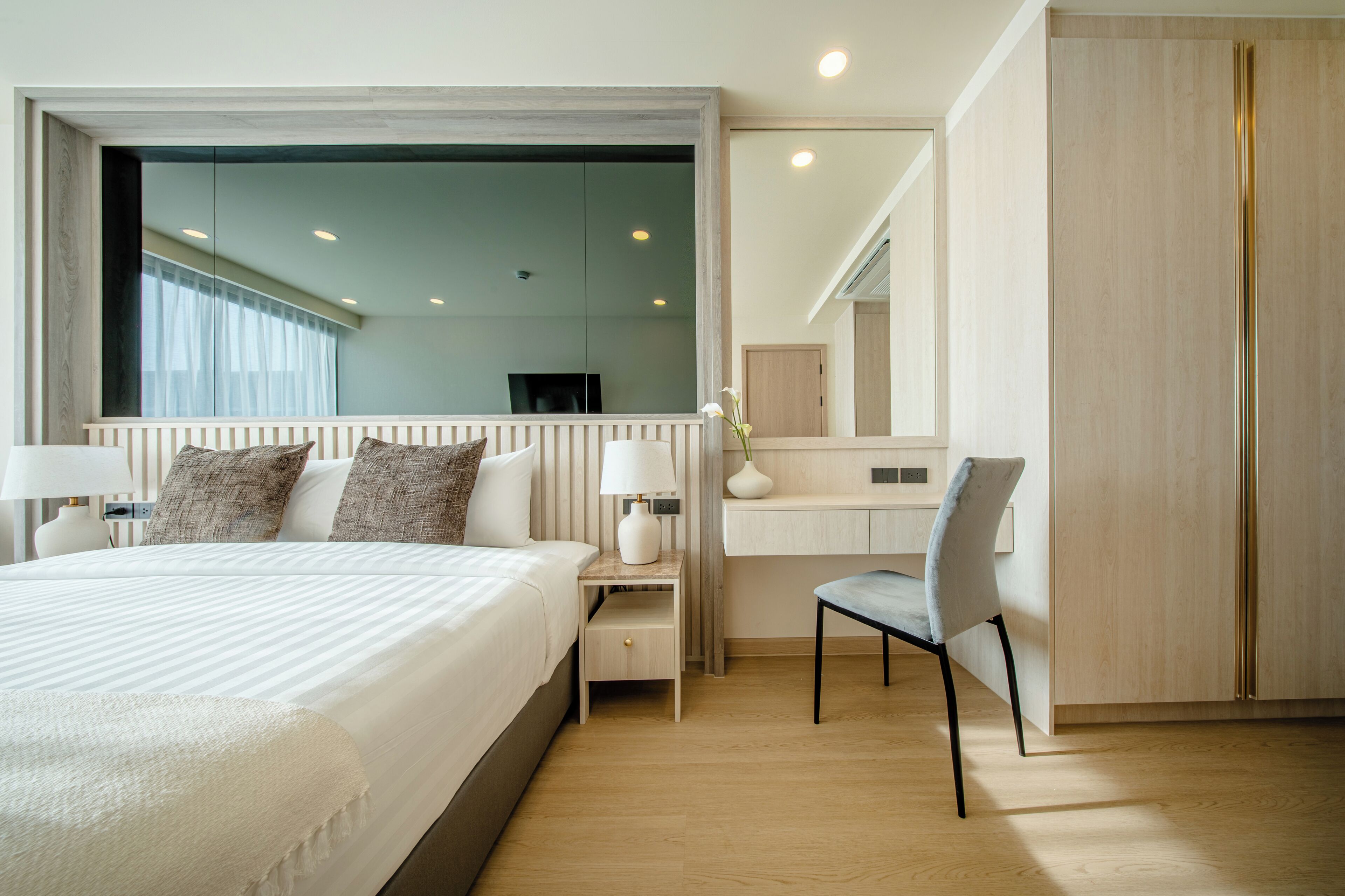A modern hotel room with a large bed, bedside tables, a chair, a vanity with a mirror, and a wardrobe.
