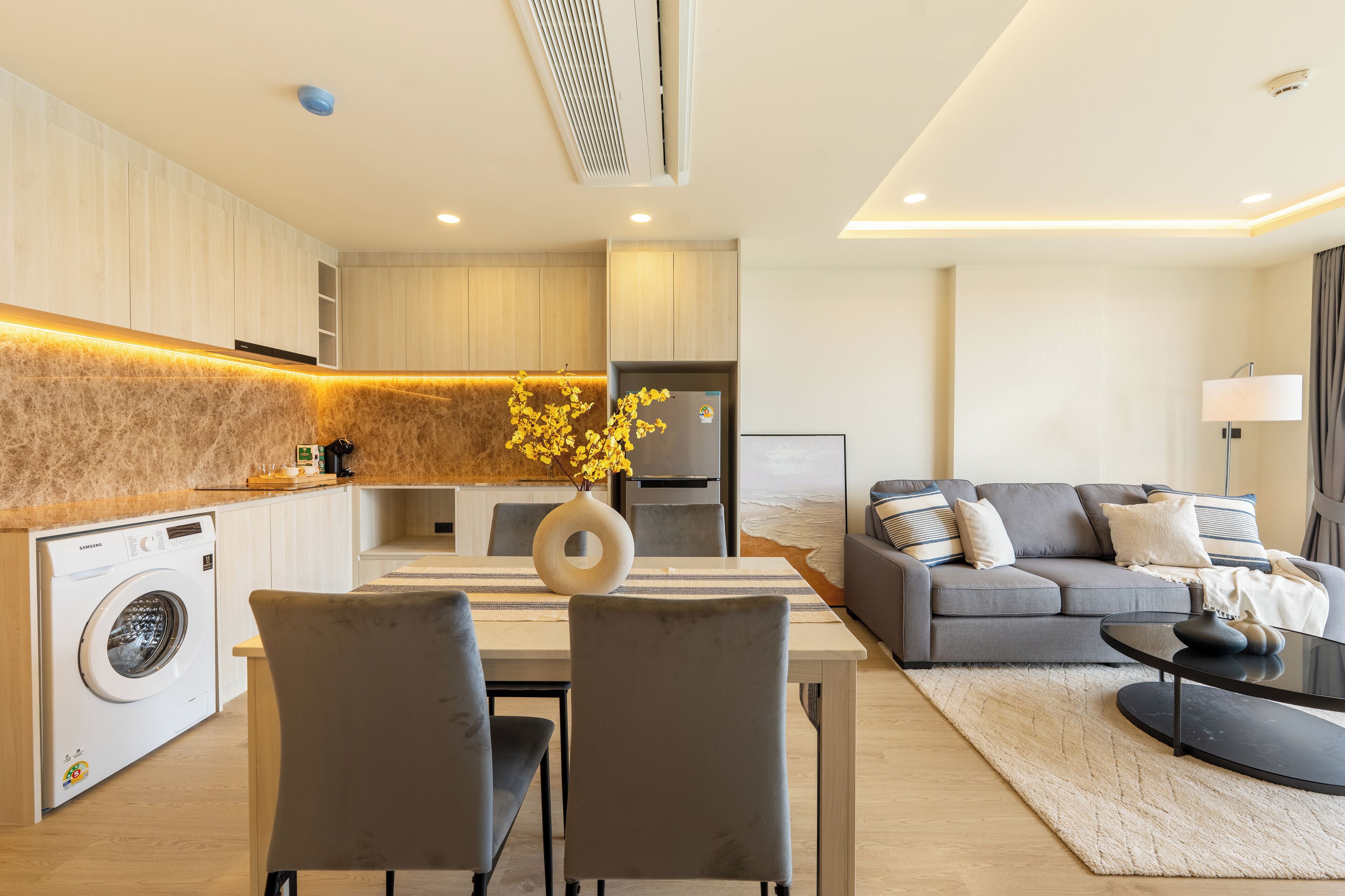 Una sala de estar moderna con un sofá gris, una mesa de comedor con dos sillas y una zona de cocina con lavadora.