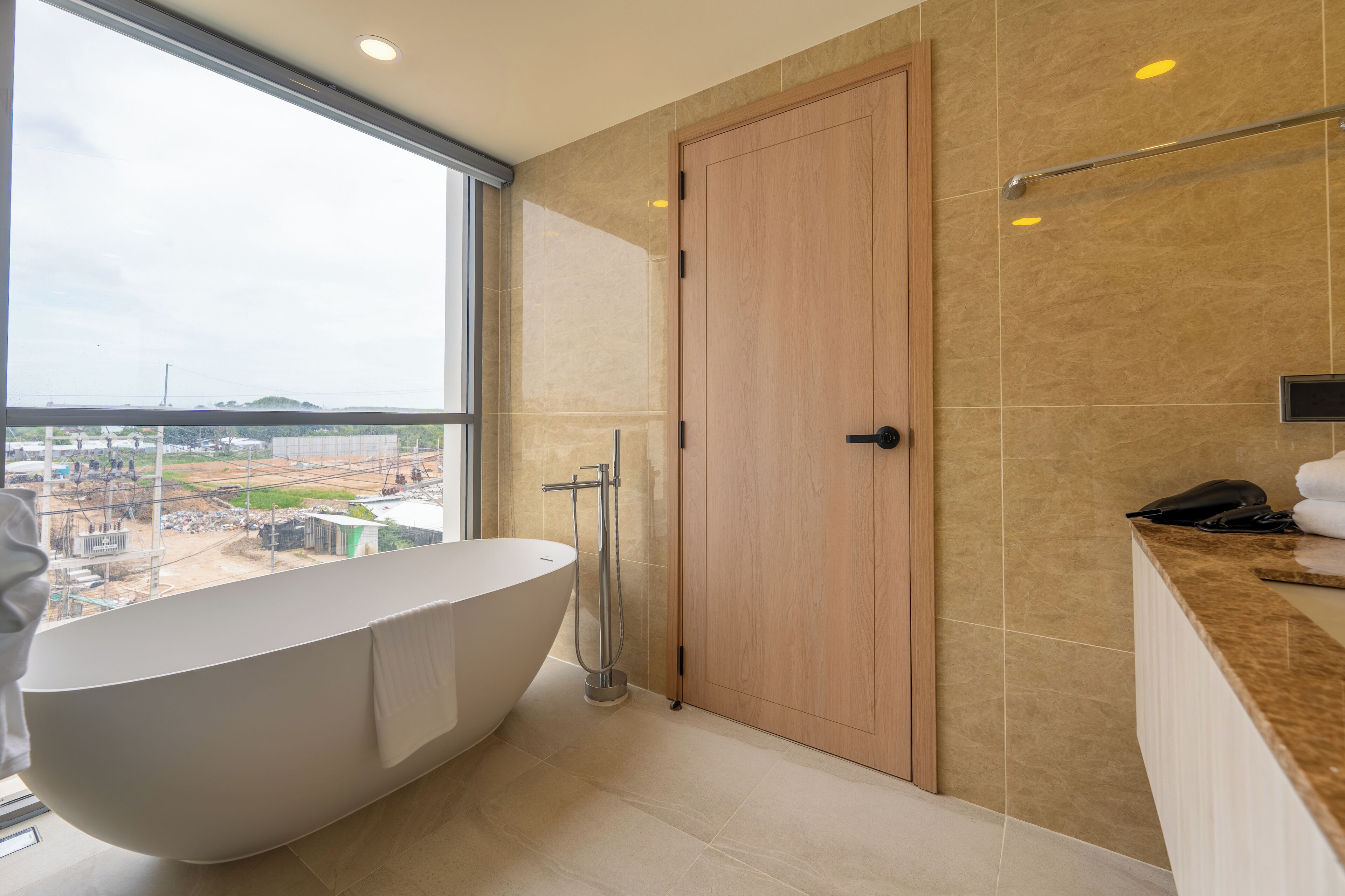 Een moderne badkamer met een vrijstaand bad, een houten deur en uitzicht op een bouwterrein door een groot raam.
