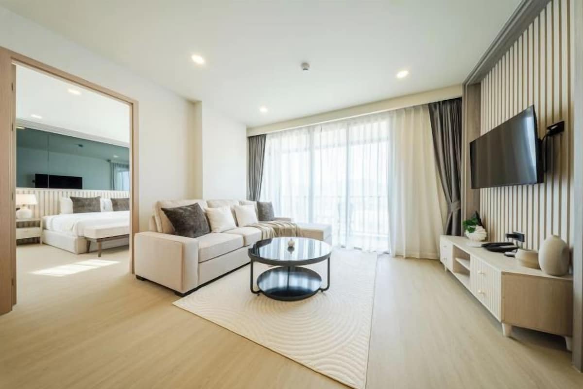 A modern hotel room with a bed, sofa, round coffee table, TV, and a large window with curtains.