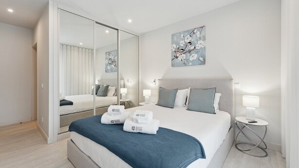 A modern hotel room with a large bed, a mirror, a painting, and a bedside table with a lamp.