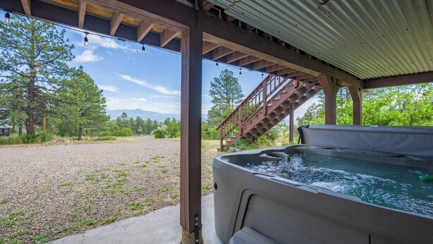 A hot tub under a covered patio with a staircase leading up to it, surrounded by a gravel area and trees.