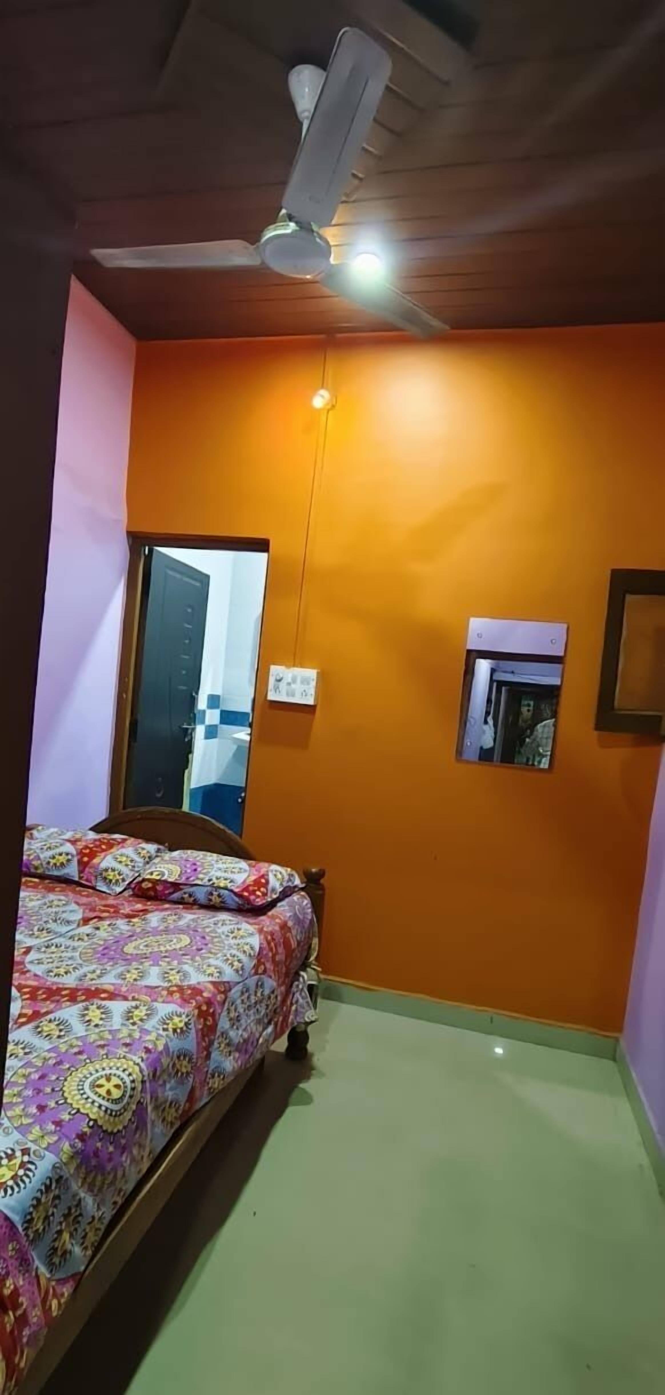 Ein Schlafzimmer mit orangefarbener Wand, einem Bett mit farbenfroher Bettdecke, einem Deckenventilator und einem kleinen Fenster.