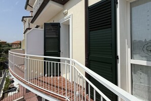 Un balcon avec une balustrade blanche, une porte avec un volet vert et une fenêtre ornée d’un rideau à motifs.