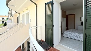 A balcony with a metal railing, a door leading to a bedroom with a bed and a nightstand, and a suitcase on the floor. - P068 - Porto Recanati, new two-room apartment with panoramic terrace (Porto Recanati)