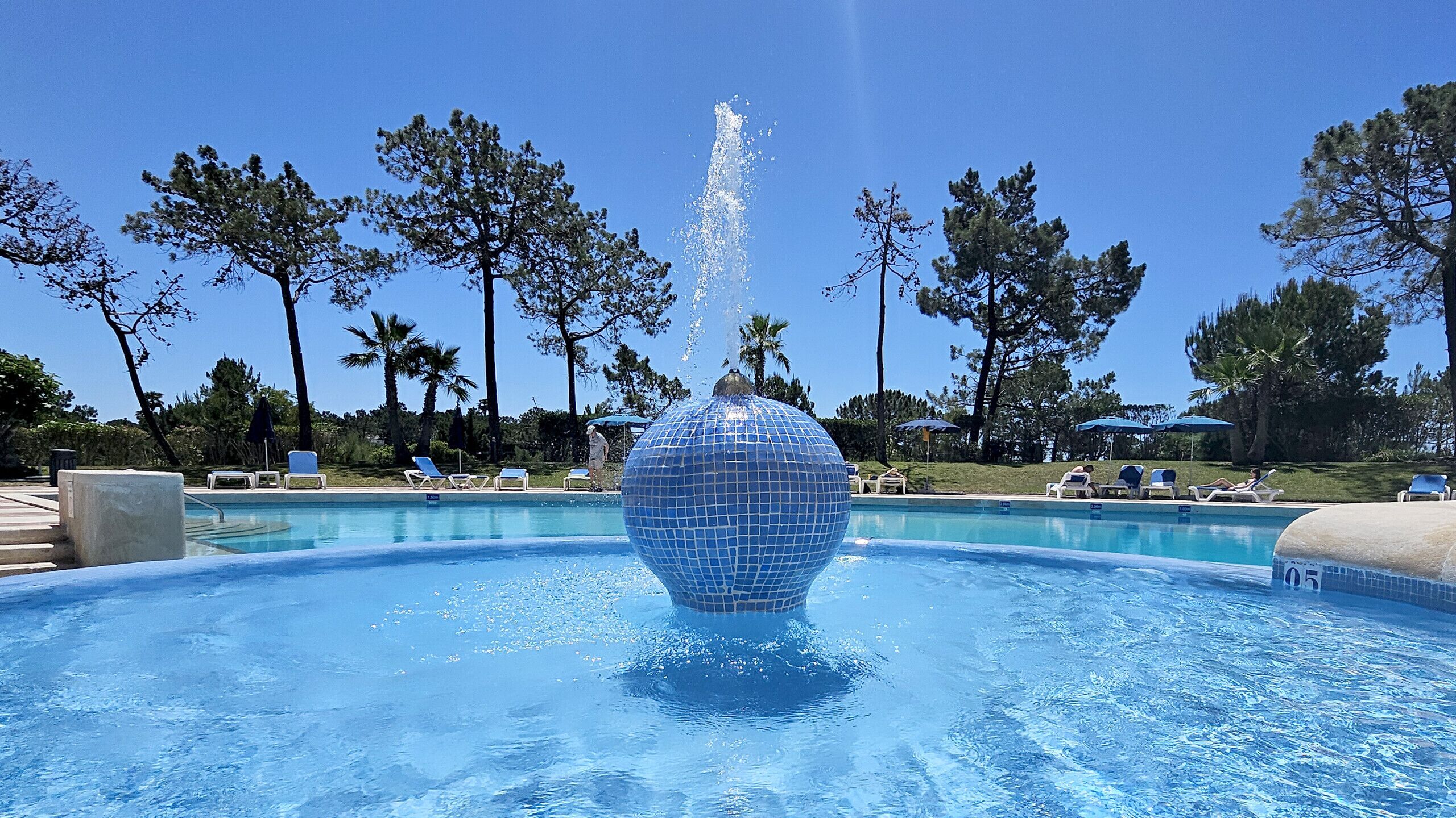 Una piscina con una fuente esférica en el centro, rodeada de sillas de descanso y palmeras.