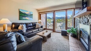 A living room with a leather sofa, a coffee table, and a fireplace. There is a balcony with a view of mountains and a lake. - Spacious Ski-In Condo in the Heart of Breckenridge! (Breckenridge)